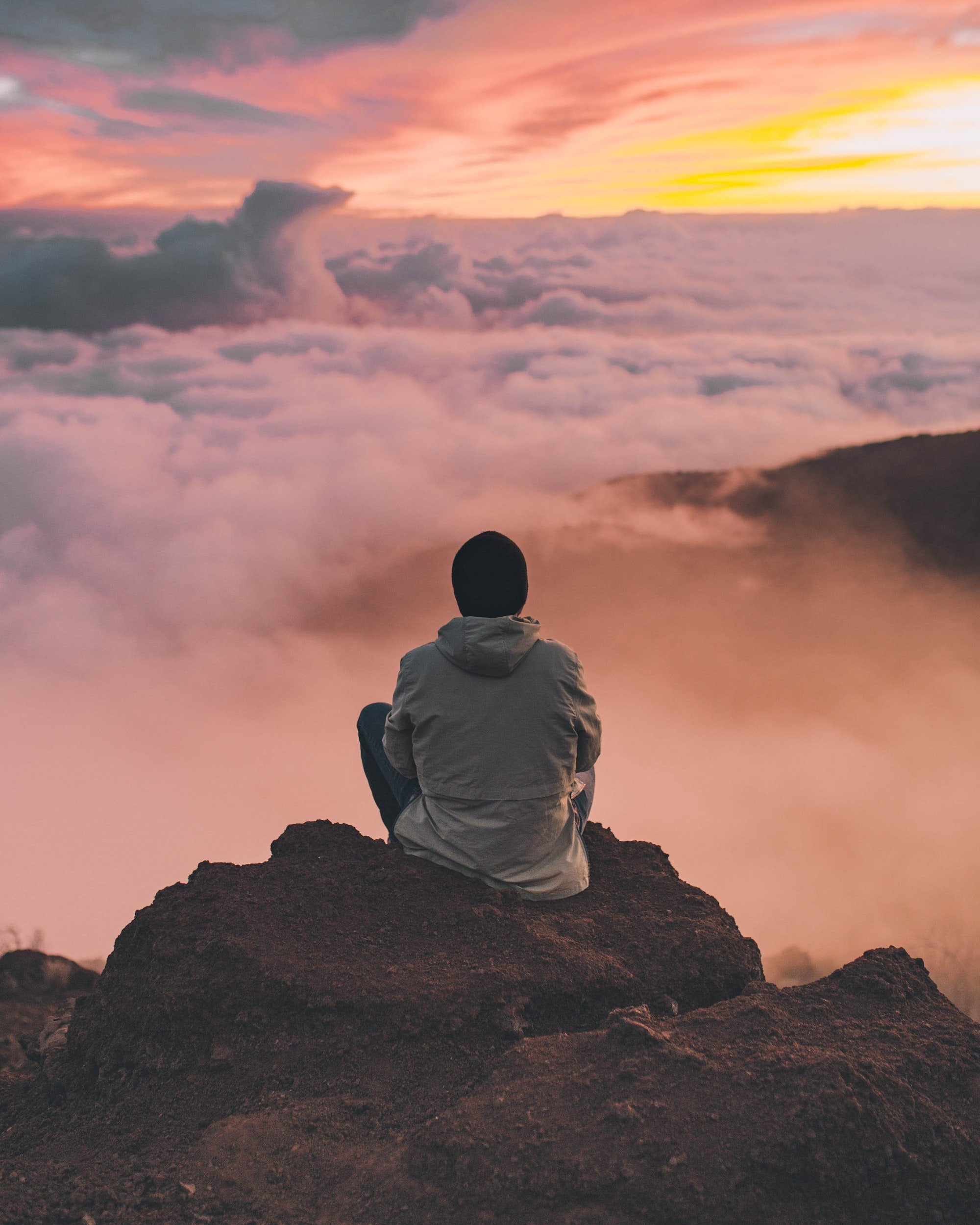 Person sitting on mountain peak overlooking clouds at sunset.
